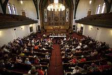 Six political candidates sit behind a short desk in a church. People fill up most of the pews in the church, and a lone microphone sits in the middle aisle.