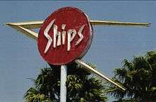 Photograph of the top of a large sign on a pole. There is a red circular section with the word "Ships" written in neon tubing. There is a larger, sideways "V" shape whose arms pass through the sign and intersect a few feet to the left of the circle. The tops of two palm trees are visible just behind the sign.