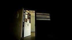 Interior view of a dark prison cell, looking out towards an open cell door, revealing a brighter exterior