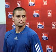 A close to bald man, wearing a blue T-shirts with a white Adidas logo, standing in front of a wall with a dusin St. Petersburg Open logos