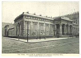 An Egyptian-Revival-style buidling on a street corner; there are doric columns on the façade to the right of the image. There are men on the street, and work is being done using cranes, on an adjacent building out of frame to the right.