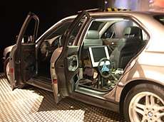 Side view of a vehicle with its doors open. Behind the left front seat can be seen a steering wheel and monitor.
