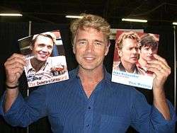 A blond-haired middle-aged man wearing a blue shirt, holding two large photographs of himself