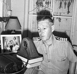Indoor half portrait of man in light-coloured military uniform speaking into radio transmitter with a family photo to his right