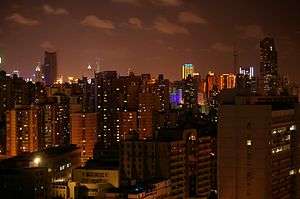 A cityscape with block-like, shadowed towers under a night sky with a bronze glow from a distant illuminated skyline in the background