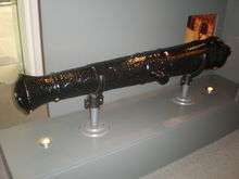 Photograph of the body of a black muzzle-loading cannon propped by two braces rest on a rectangular gray stand with two embedded little round lamps.