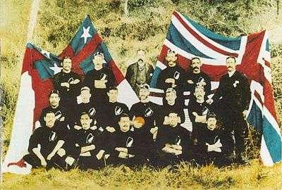 Photograph of the Native football team in their uniform of black shorts and jerseys sat in front of the flags of Great Britain and the United Tribes of New Zealand