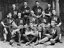 A black and white photograph of twelve men arranged in three rows (standing, sitting in chairs, and sitting on the floor) each wearing a dark baseball uniform with a white "N" on the chest; some are holding dark and white striped caps