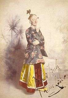 Colourised photograph a man standing in a yellow skirt and patterned silk top, with his hair worn in a comical topknot and chin resting on one finger, looking sideways towards the viewer