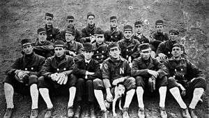 A black and white photograph of seventeen men sitting in three rows on the ground wearing dark baseball uniforms and caps with high white socks; one is dressed in a suit and another is holding a dog
