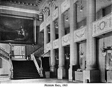Black-and-white photo of a two-story interior room. There are large columns on the right-hand wall and a wide staircase on the back wall. A large painting hangs above the staircase landing.
