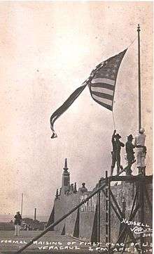photograph of a walled fort with three Marines raising a U.S. flag over it