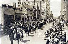 Hundreds of men on horseback march down a city street as people observe from the sidewalks and rooftops.