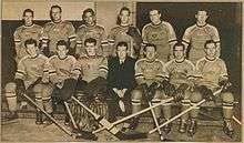 A black and white photo of ice hockey players sitting in two rows on a bench, with their coach in the middle front row.