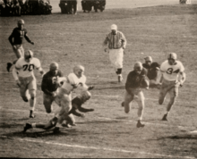 A man in a black jersey about to run a touchdown against several players in white jerseys