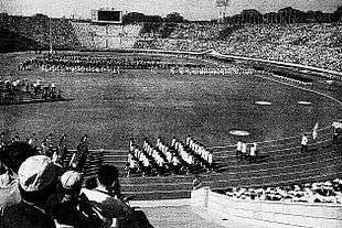 A black-and-white photograph of a stadium with athletes marching at the track