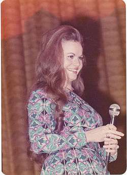 A young woman with long brown hair wearing a dress with an intricate multi-coloured pattern and holding a microphone