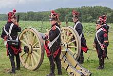 Photo of four reenactors dressed as French artillerymen in dark blue uniforms. They stand near a replica of an early 1800s cannon.