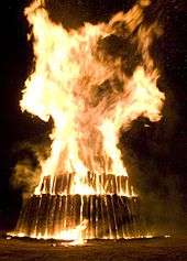 Large flames engulf the top layer of a circular three-tier structure of logs; the logs are vertical to the ground.