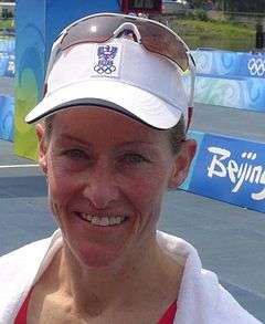 A blue-eyed smiling woman wearing a white towel around her shoulders and a white cap with a pair of white-framed sporting sunglasses on top of it. The cap bears the Austrian Olympic committee logo.