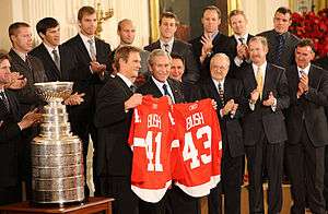 In the front row, Nicklas Lidstrom holds a Red Wings jersey with the last name "BUSH" and the number "41" while George W. Bush holds a Red Wings jersey with the last name "BUSH" and the number "43". To Lidstrom's right is the Stanley Cup. Second row, left to right: Mike Babcock, Ken Holland, Jim Devellano, Jim Nill, unknown man.  Third row, left to right: Kris Draper, Pavel Datsyuk, Mikael Samuelsson, Brian Rafalski, Derek Meech, Kirk Maltby, Chris Osgood, Aaron Downey.