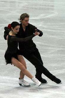 A man and woman ice dancing; the woman, on the left, has dark hair pulled back tightly with a rose tie and wearing a short black dress, and the man is wearing an open-collar black shirt and black trousers