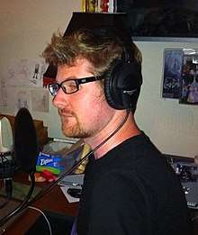 The image depicts a man wearing glasses and headphones (pictured in 2010).