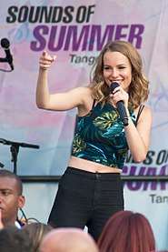 Young, blonde-haired woman gesturing towards her audience while smiling and singing into a hand-held microphone.  She is outfitted in a floral-themed tank top and black pants; her midriff is exposed.