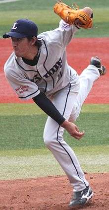 Takayuki Kishi wearing a gray baseball uniform and blue baseball cap in the process of pitching a baseball