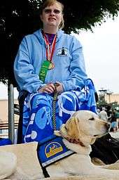 A Yellow Labrador Service Dog in a blue service dog vest with a yellow Logo lies in the foreground of the photograph. A woman sits behind him, in a wheelchair, with a blue shirt, long flowing skirt, sunglasses, and has an award medal hanging around her neck.