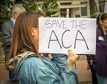 A hand-lettered sign saying "Save the ACA" held up by a woman standing in profile to the left. It blocks her facial features from view