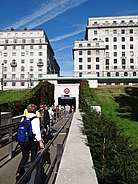 Entrance from Green Park, a straight footpath alongside a stone wall topped with a hedge leads towards a square entrance set into the side of a slope. Two large buildings fill the skyline.