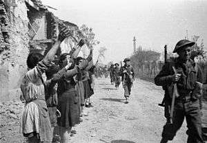 A black and white photograph of men wearing military uniforms marching along a road with civilians in front of their houses cheering them on
