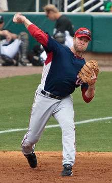 A man wearing a navy-blue baseball jersey and white baseball pants throws a baseball with his right hand