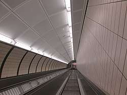 Looking down a bank of two escalators in a semi-elliptical passage lit by fluorescent light strips above, generally white with some yellow paneling on the walls. There are no people on the escalator. The wall on the right-hand side is flat and contains a vertical tiling pattern, while the wall on the left-hand side is curved with tiles patterned diagonally and parallel to the escalator.