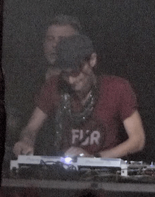 A DJ with a red shirt and black cap performs live on a stage, with former Underworld member Darren Emerson standing behind him