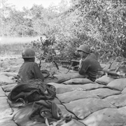 Two soldiers manning a machine-gun in a heavily sandbagged pit observe the perimeter of their defensive position through the vegetation