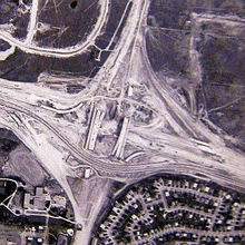 A bird's-eye view of a large highway interchange under construction. Several bridges are complete, but nothing is paved, aside from one highway crossing horizontally, which detours between the bridges.