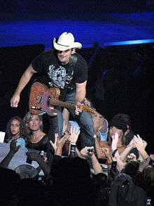 A man in  a dark t-shirt, jeans and cowboy hat playing the guitar on a stage in front of an audience