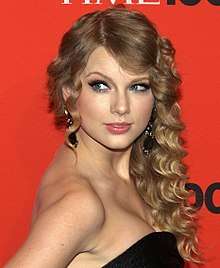 Taylor Swift stands in a Time press area, wearing a black, strapless dress and curled hair