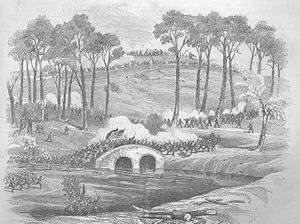 A 19th century engraving of a landscape in which a military action is taking place. Two opposing forces of soldiers battle over a stone bridge. Two flags are carried by soldiers at the center of the bridge. Troops approach from a high hill in the background and white smoke rises on the hillside.