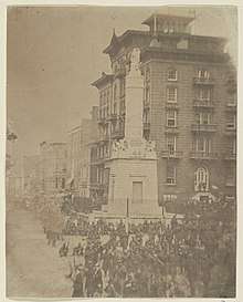 An antique photograph depicting a city square with a stone monument and a large number of soldiers at rest