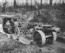 A large field gun and limber being towed along a sunken road through a battle-scarred forest by a Holt tractor. The gun has large fabricated steel-spoked wheels, with a wide tread to cope with mud.