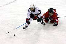 A female ice hockey player, wearing white, plays the puck as an opposing player in red falls down on the ice trying to stop her.