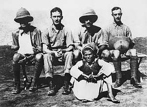 Four men in khaki military uniforms, two wearing pith helmets (one of whom has his arm in a sling), seated behind a boy