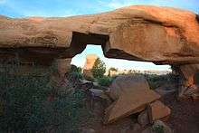 Mano Arch—much thicker than Metate Arch—with several boulders lying under it and a large notch missing from the bottom of the arch. A distant hoodoo appears framed inside the arch's notch.