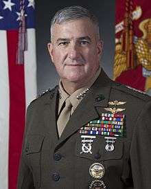A color image of Glenn Walters, a white male in his Marine Corps Service A uniform. He is not wearing a hat, ribbons are visible. The Marine Corps flag and United States flag are visible in the background.
