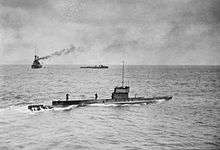 A submarine moves across the surface of the ocean, while two warships can be seen in the background.