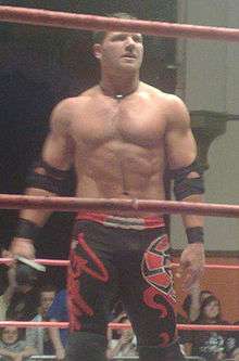 A white male wearing black and red wrestling gear standing in a wrestling ring.