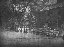 A line of soldiers in tropical uniforms stand in front of a hut in a jungle clearing. In front of them stand a group of five men facing towards the camera.